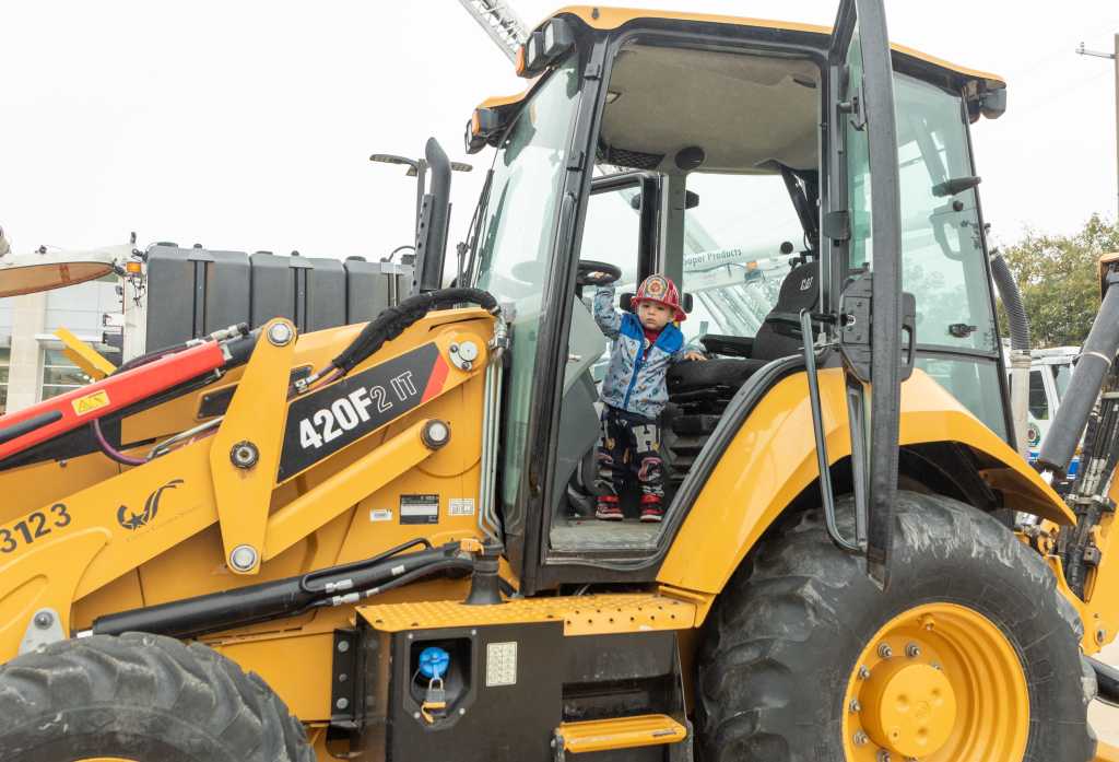 A young child wearing a fireman's hat is sitting in the driver's seat of a large yellow construction vehicle, appearing excited and engaged.