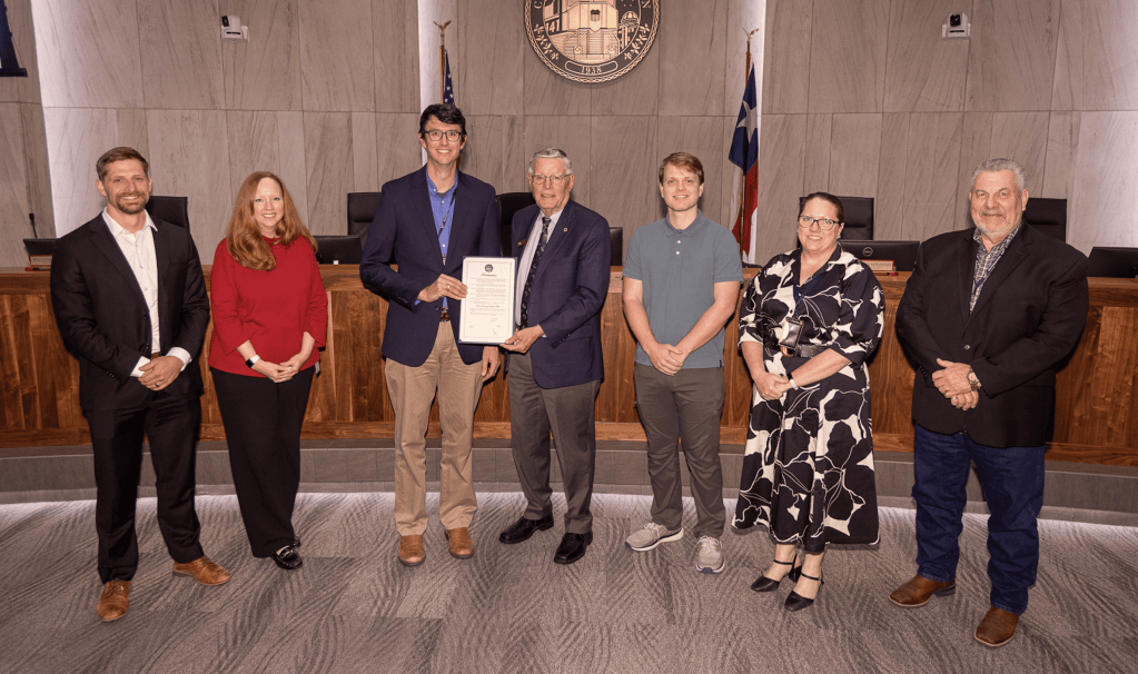 A group of six individuals stands in a government meeting room, with a dignitary holding a proclamation in the center. They are dressed in formal and business casual attire, with a backdrop featuring a Texas state flag and a seal.