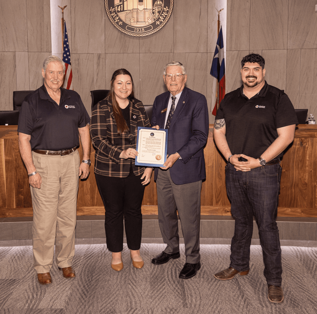 Four people standing in a formal setting, presenting a certificate. Two individuals on the left in professional attire, one holding a document, and two individuals on the right in black shirts, smiling. Behind them is a wooden backdrop with flags.