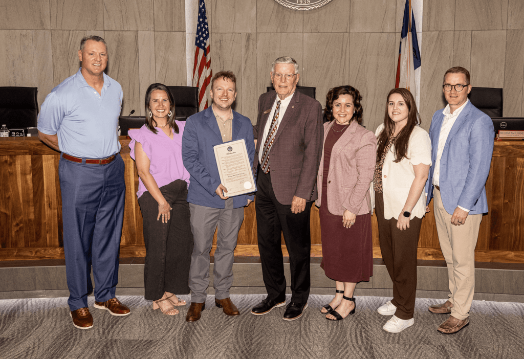 Group of seven people posing in a governmental setting, with one person holding a framed certificate, flanked by flags and a wooden panel background.