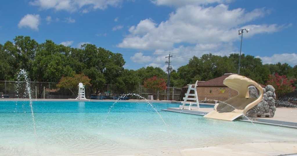 Outdoor swimming pool with a water slide, surrounded by green trees and a clear blue sky.