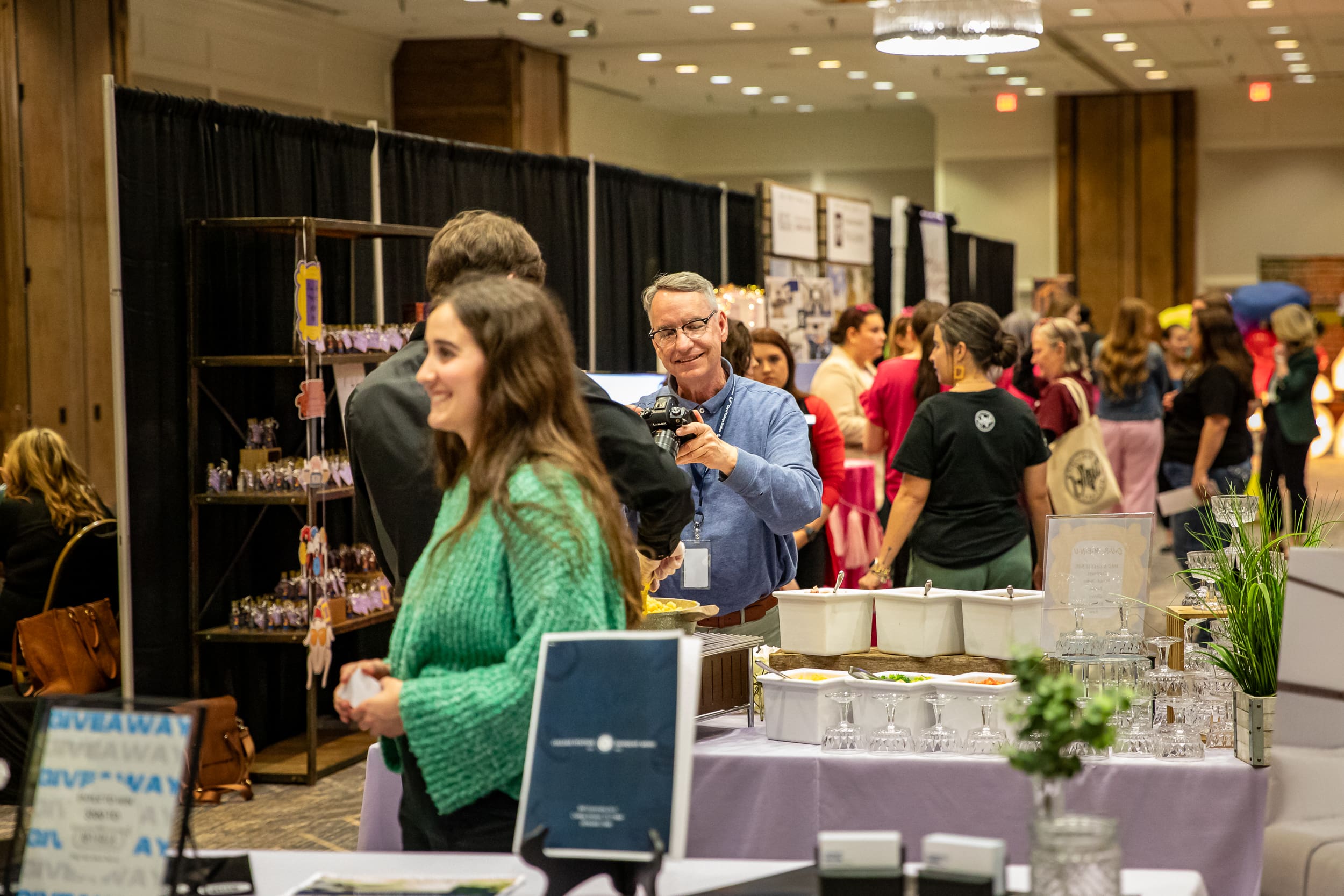 A busy event scene featuring people engaging with vendors and displays. A man is taking a photo, while a woman in a green sweater smiles, and a table has food and promotional materials.