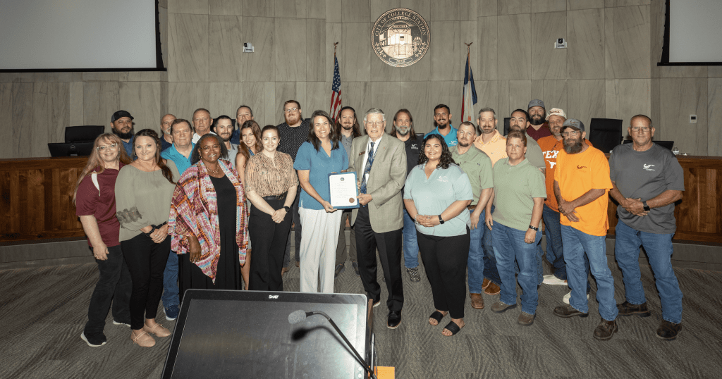 Group photo of a diverse gathering of individuals, including men and women, standing together in a formal setting with a podium. Some individuals are holding certificates, and a seal is displayed on the wall in the background.