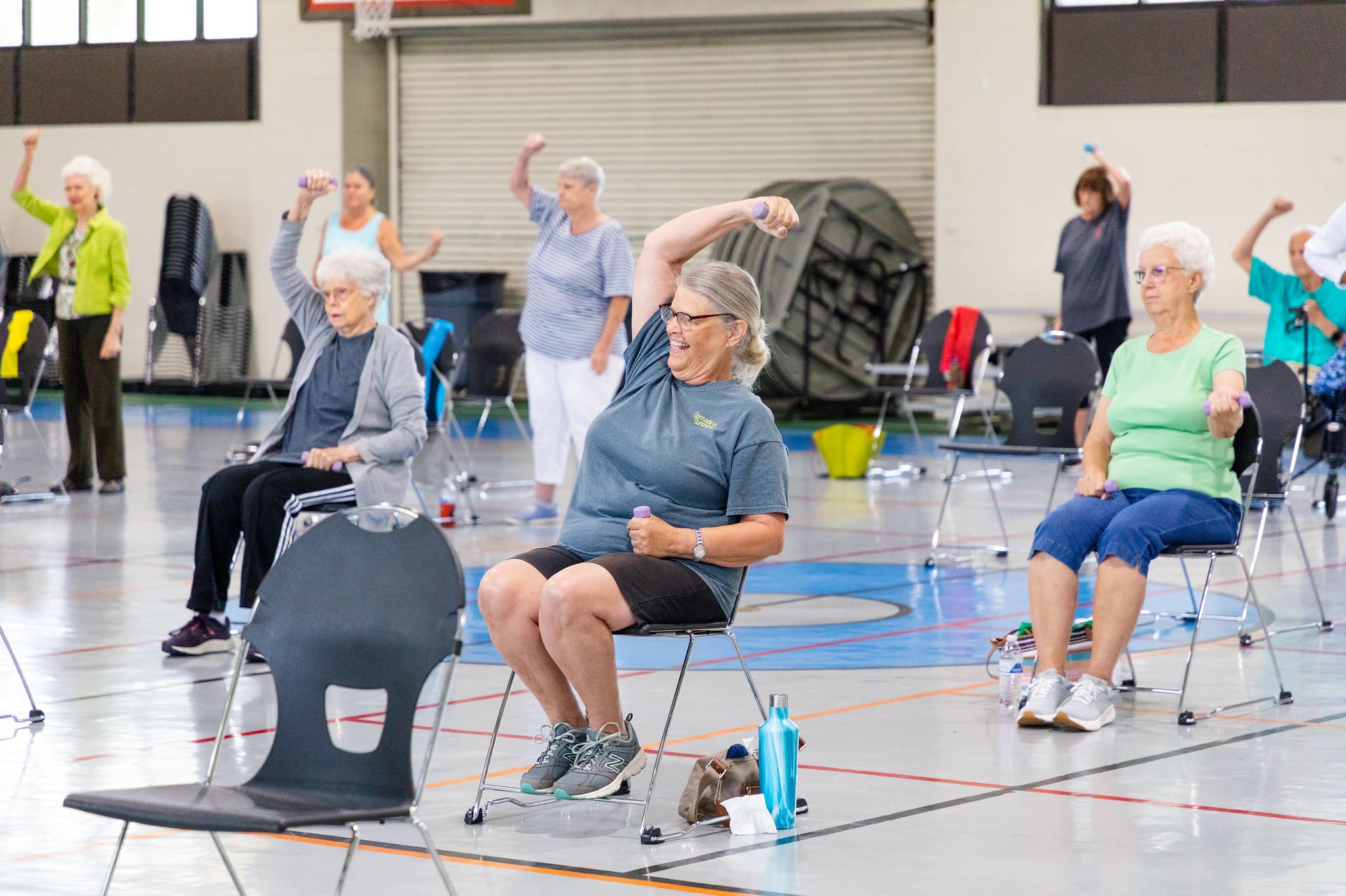 Senior adult sits on a chair in a gym doing aerobics. The person appears to be smiling.